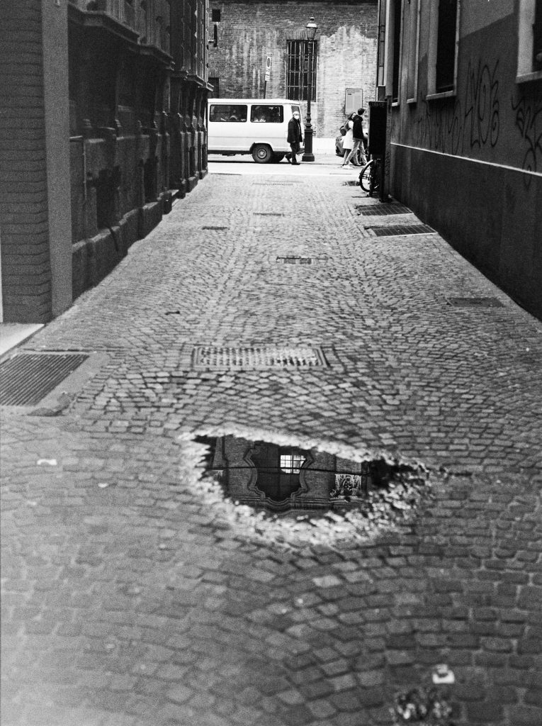 Black and white photograph of an alleyway in Parma, with a cobbled street and a puddle reflecting a sculpted window. ©Sarah Beeusaert